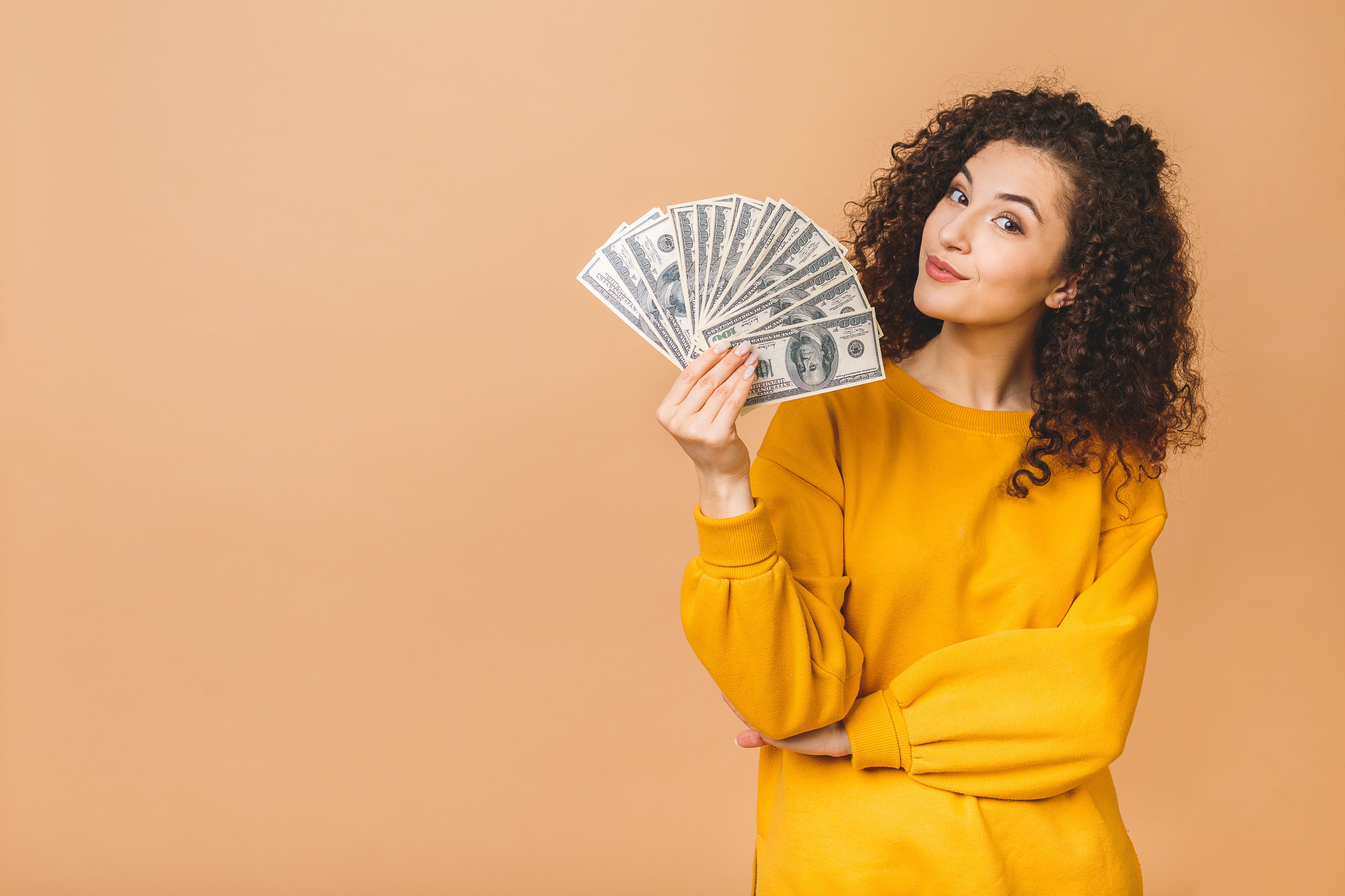 Portrait of a cheerful young woman holding money banknotes and celebrating isolated over beige background.