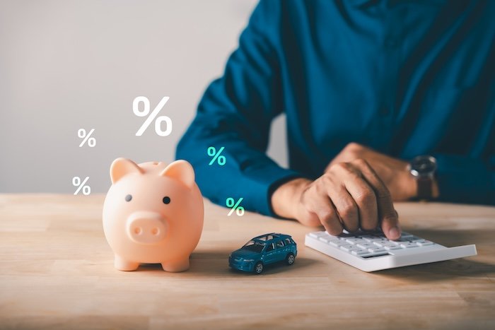 Man calculates car loan with piggy bank, saving money for auto insurance payments, bank offers safety for tax and budget planning, considering interest rates for vehicle