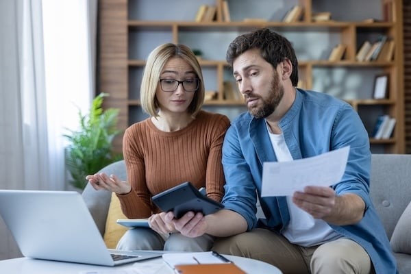 Young couple sitting on a sofa at home, managing household finances, calculating bills, reviewing papers, and experiencing stress about financial decisions and debt 