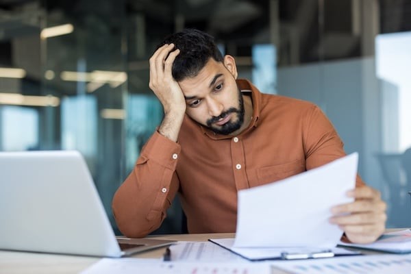 Young male employee feeling tired and frustrated while looking at financial papers at his office desk, dealing with a deadline, stress, and professional burnout