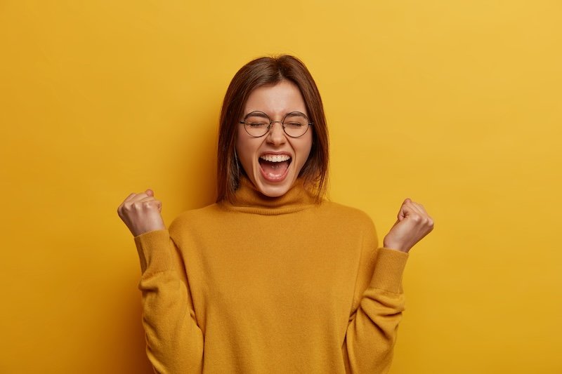 Overjoyed brunette young lady clenches two fists, celebrates success and exclaims with joy