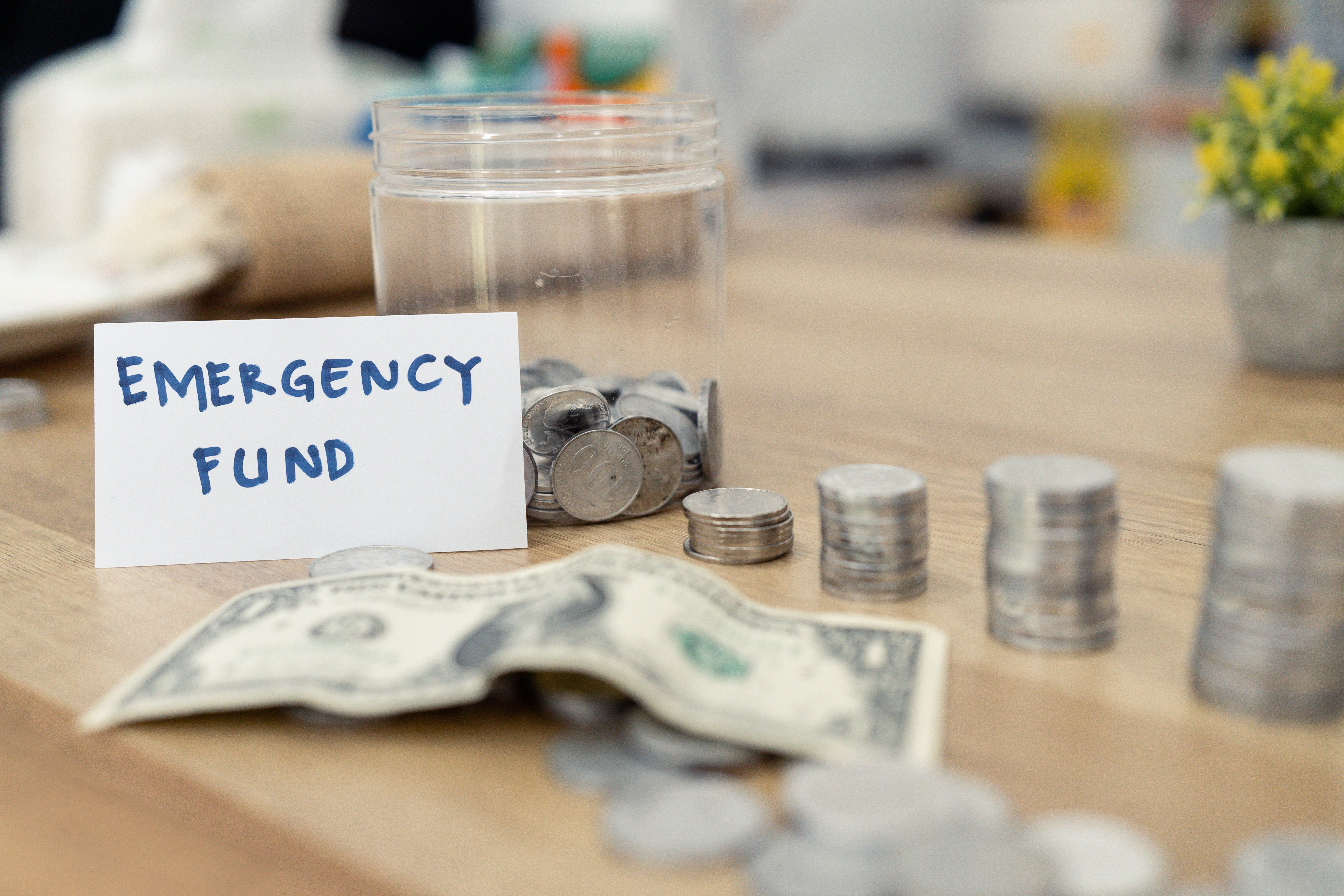Clear jar marked emergency fund with stacked coins, folded dollar bills, and loose change on wooden table