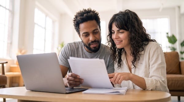 Attentive diverse young couple reviewing financial paperwork and monthly budget planner on laptop computer while managing household expenses, bills and family savings in bright living room.