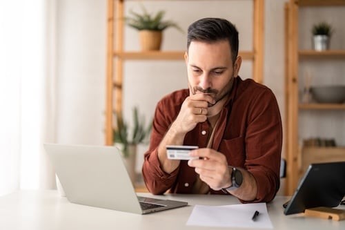 Serious business man sitting at table in home office, holding and looking at credit card, worried about debt and credit card deficit, considering a financial loan from bank