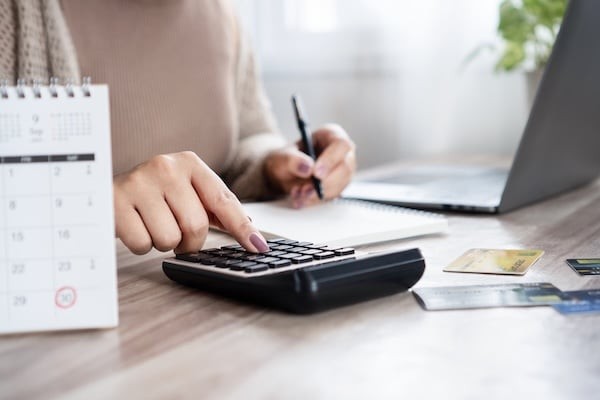 Woman calculating monthly debt payments, writing down numbers with a calendar with marked due dates, using a calculator with credit card on the desk