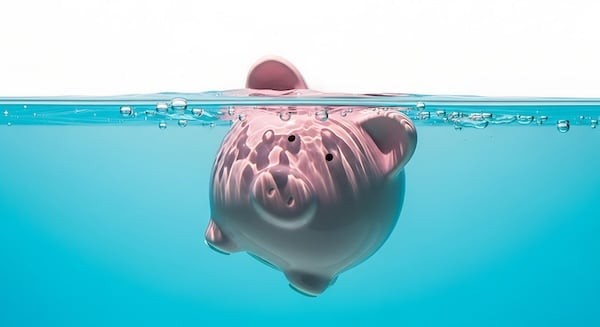 A pink piggy bank submerged in clear blue water, with only its back and snout visible above the surface, symbolizing financial trouble.