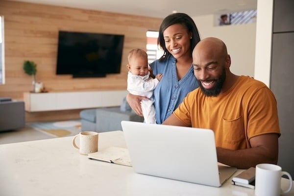 Dad sitting at the counter on a laptop while mother holding baby stands behind him smiling at the screen