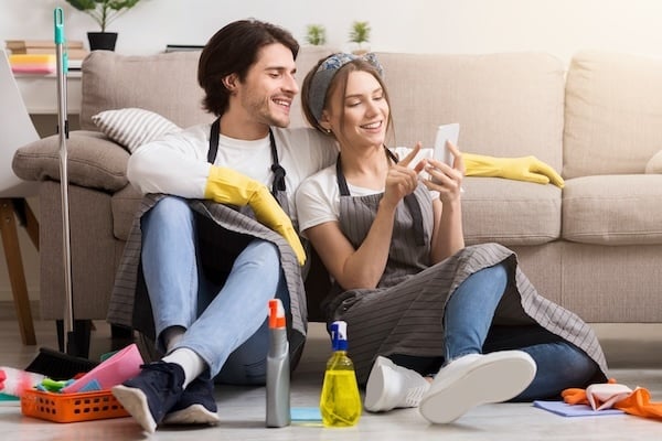 Smiling young couple relaxing with smartphone after spring-clean apartment, sitting on floor with lots of detergents and cleaning supplies