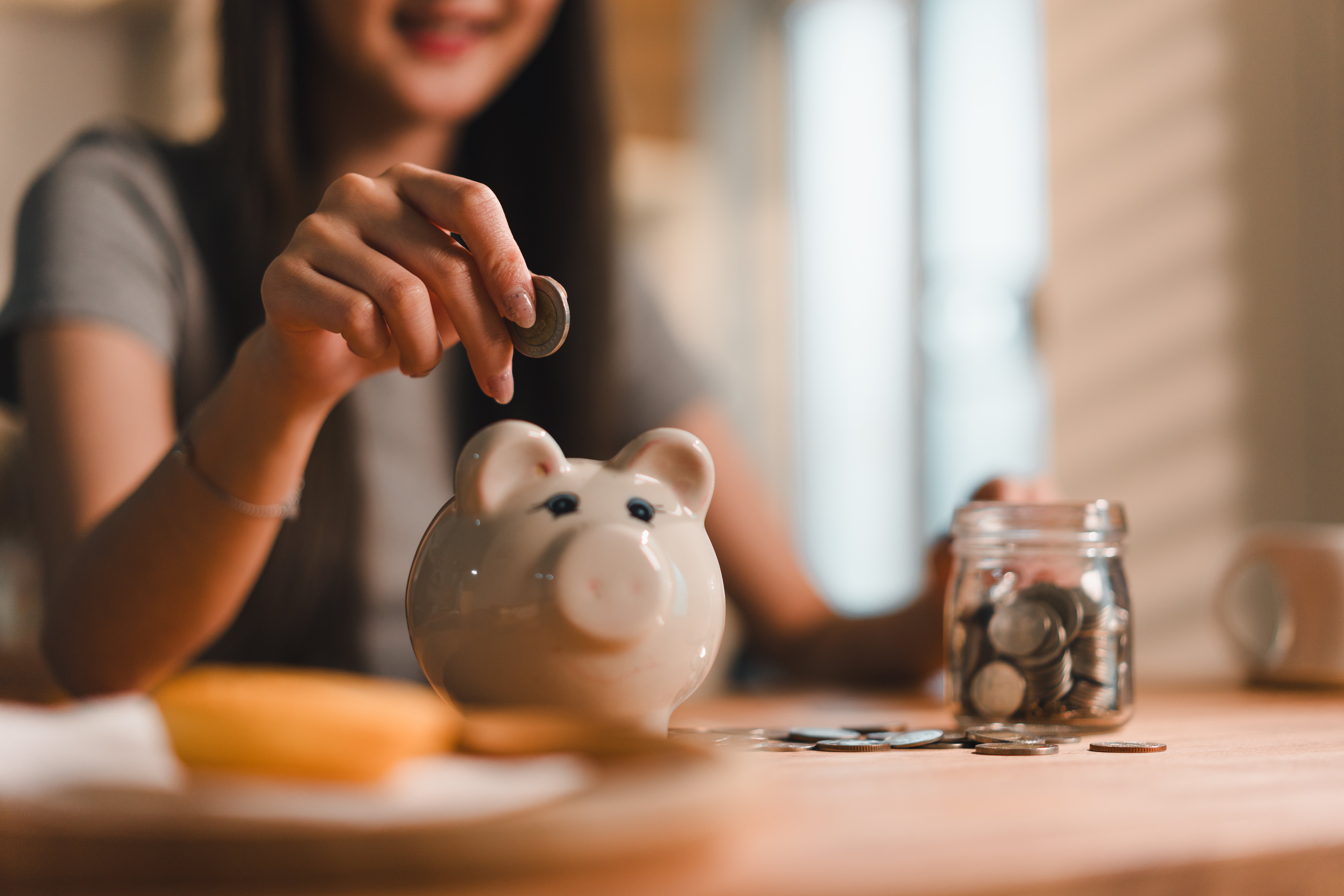 woman placing coin into piggy bank while surrounded by coins and jar filled with more coins Adobe stock image